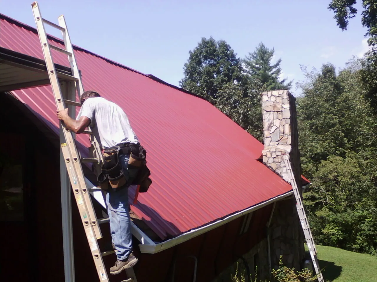Roofer on a ladder beside a red metal roof and stone chimney — Soffit & Fascia Repair by Roofing Brothers in Ringoes